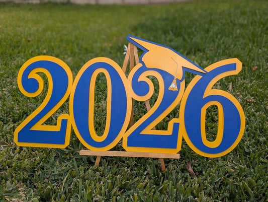 A wooden Graduation sign spelling '2026' in bold blue letters with yellow outlines, topped with a small yellow graduation cap, displayed on a green grass lawn.